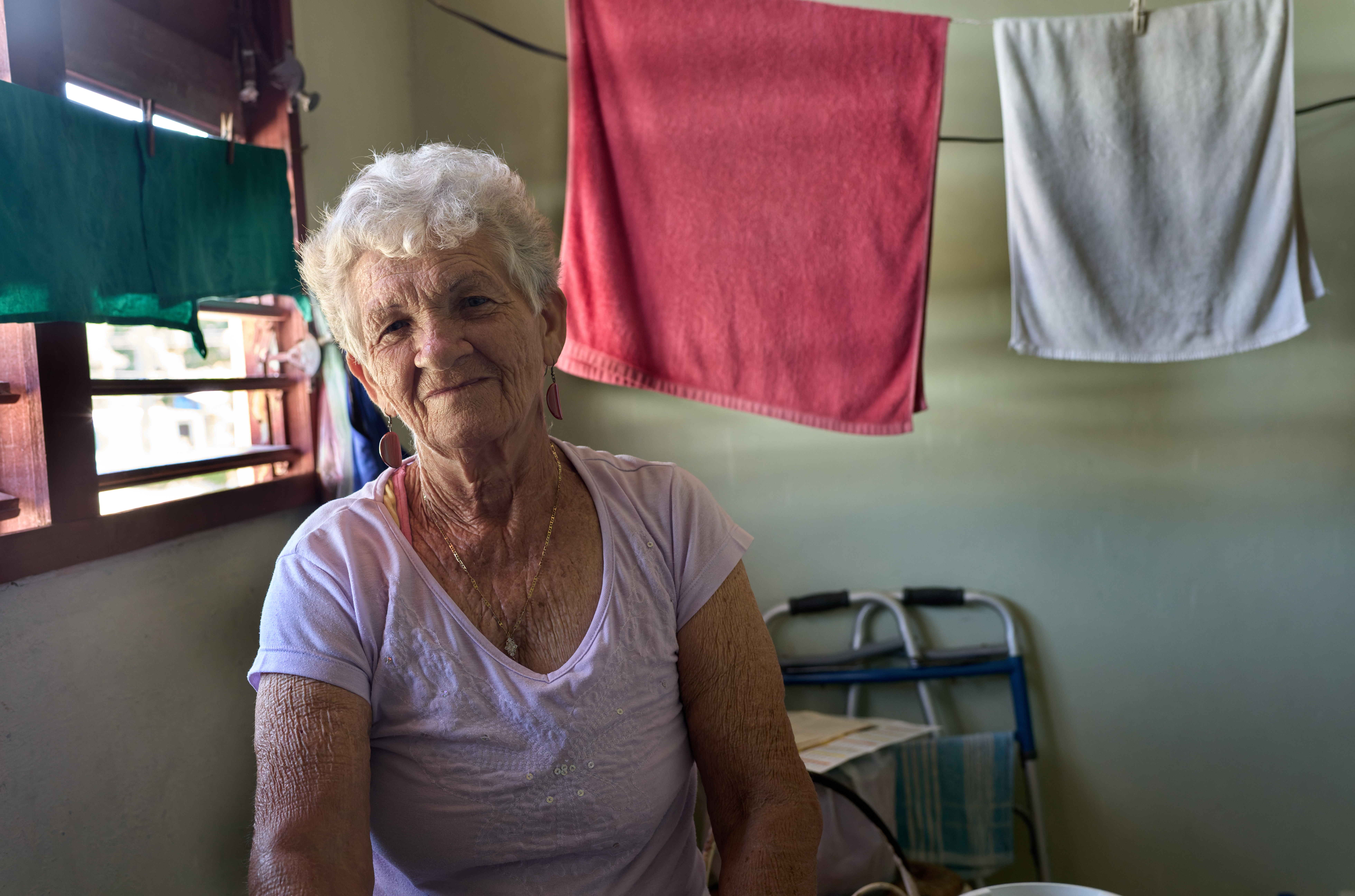 Caridad at Home:    Surrounded by drying laundry and quiet light in the dining area, Caridad sits beside her husband’s walker in their Central Havana apartment. She now cares full-time for her ailing partner.