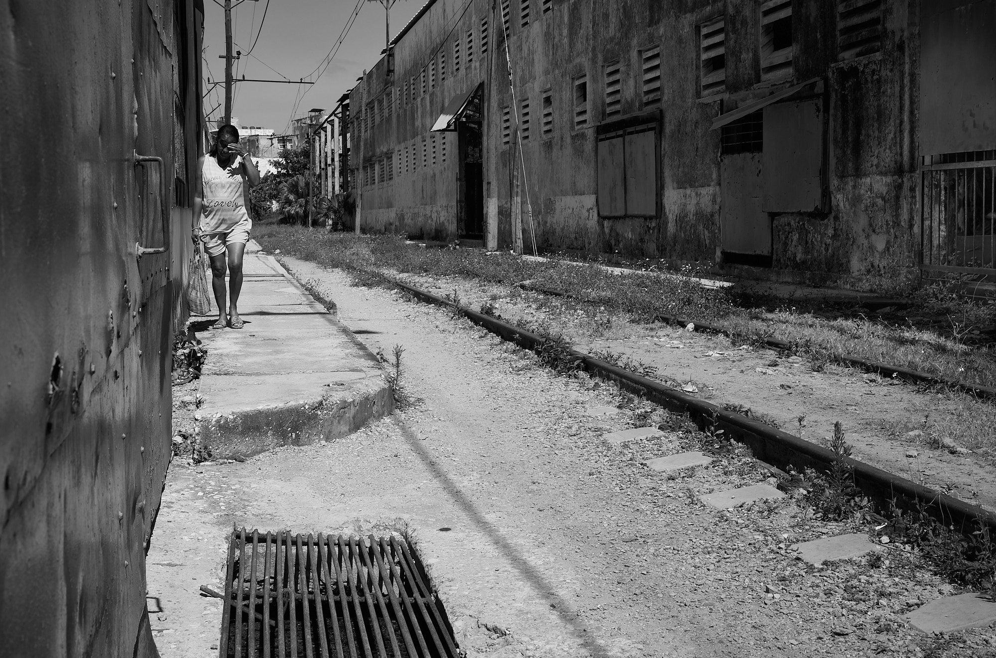 Groceries in hand, a woman strolls through the same industrial zone where I photographed the workers behind the cage at the far right. Just steps away, men shape sand molds for marine propellers — bare-handed, with no masks or gloves. The ground outside the caged area is slick with decades of oil and residue, carrying toxic materials beyond the workshop itself. There are no signs, no warnings, no fences.