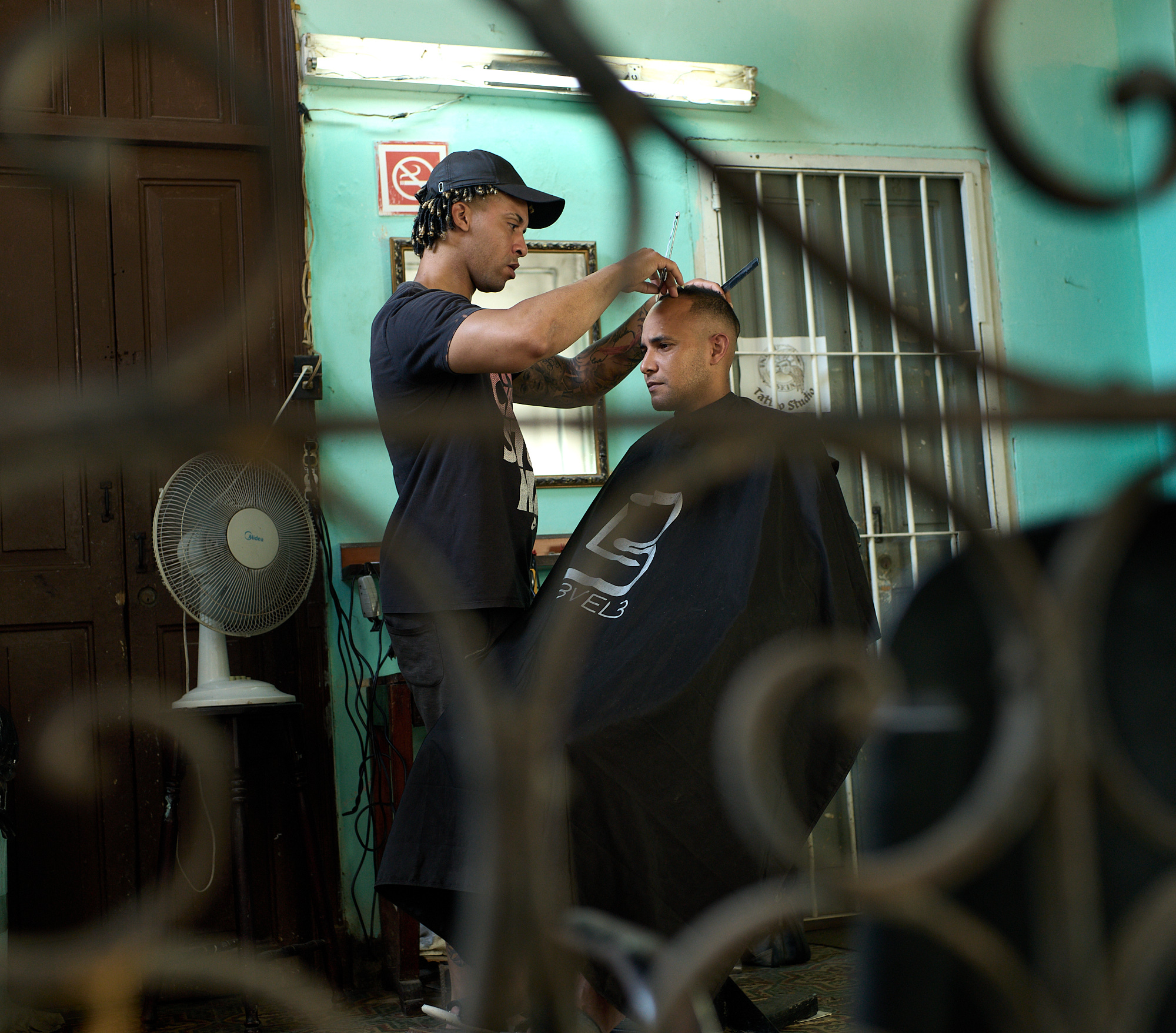 Outside his Havana apartment, the barber I interviewed works where neighbors come and go, his chair and mirror tucked beneath the shared entryway. For thirteen years, this small space has been both his shop and his livelihood.