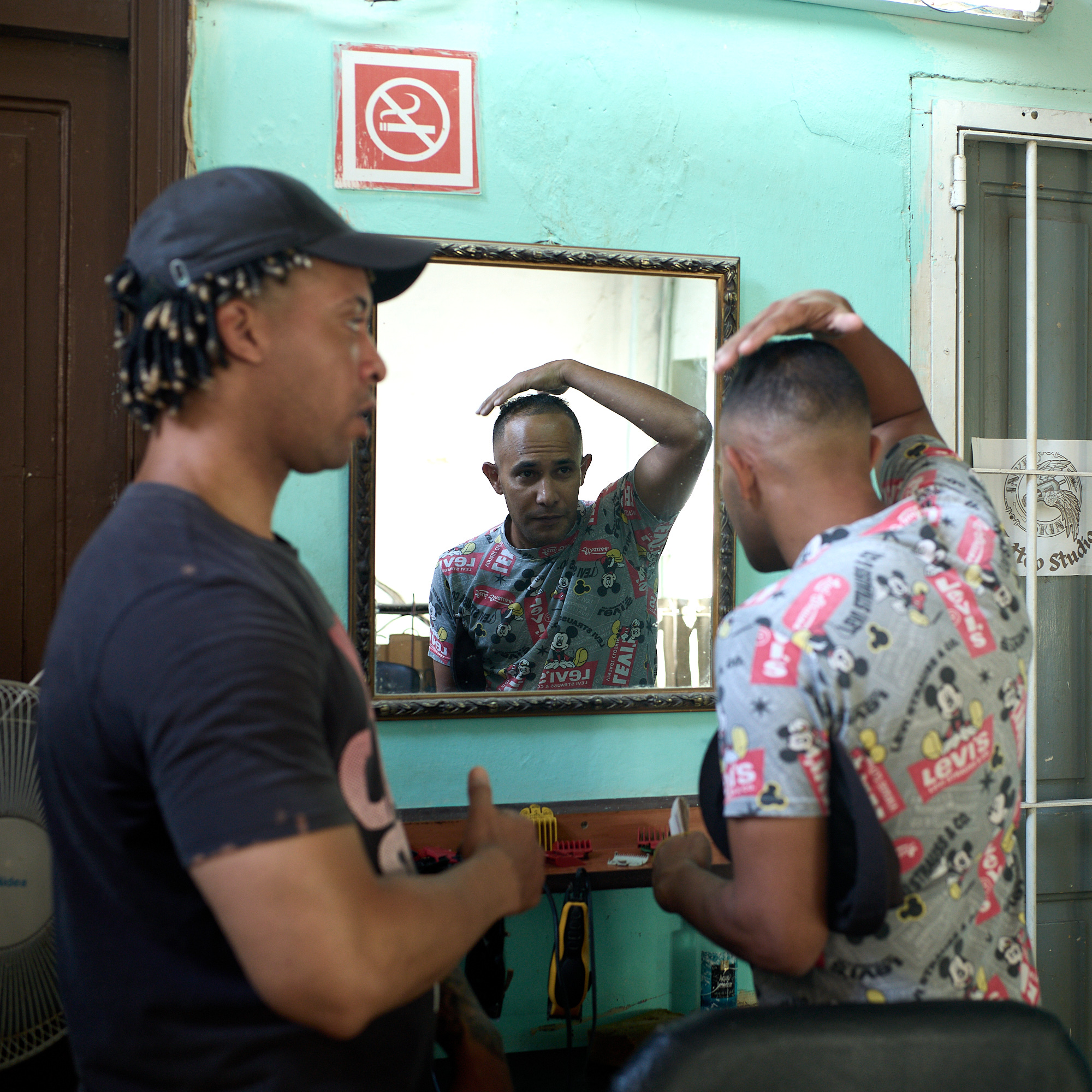 In the reflection of a small mirror, a client inspects his fresh cut while the barber waits, clippers in hand. It’s a moment of quiet satisfaction, a simple exchange of trust and pride in a makeshift Havana barbershop.