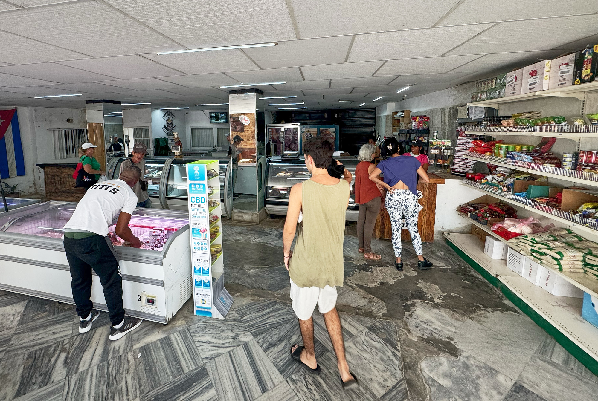 Customers quietly move slowly through Charcutería y Pescadería Titi’s, a state-affiliated butcher shop in Havana where supplies arrive sporadically and refrigeration is unreliable. The Cuban flag hangs above near-empty coolers, a quiet emblem of endurance in a shrinking economy.