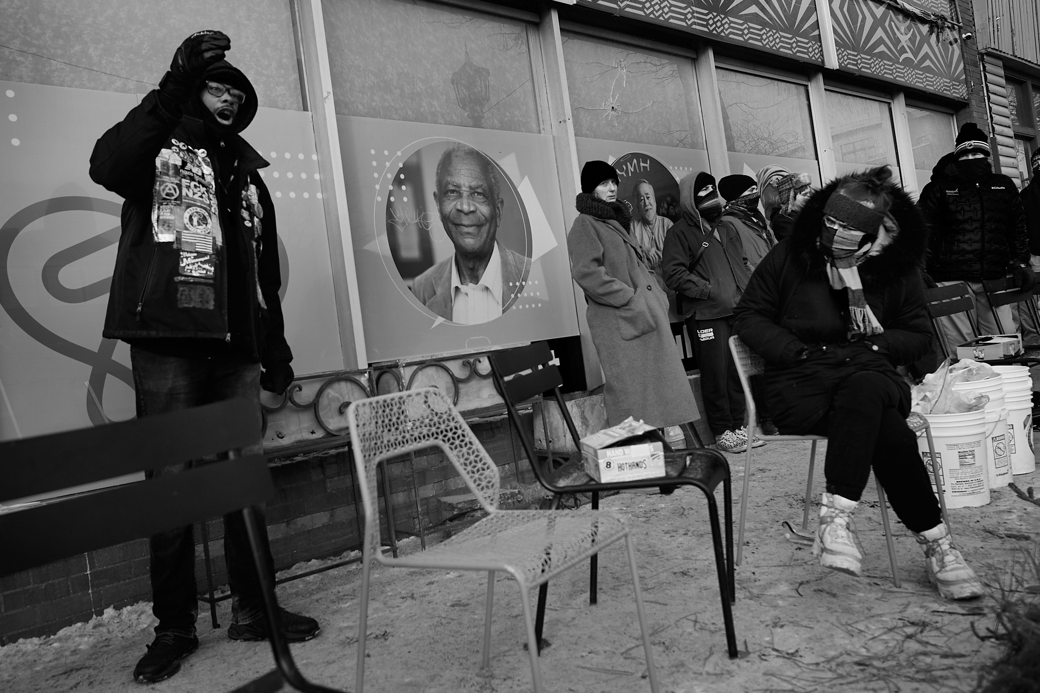 A man speaks forcefully during a protest as a seated woman listens with visible skepticism, as he criticizes what he described as documenting injustice without participating in meaningful change, highlighting internal tensions over action, accountability, and the role of observers within the movement.
