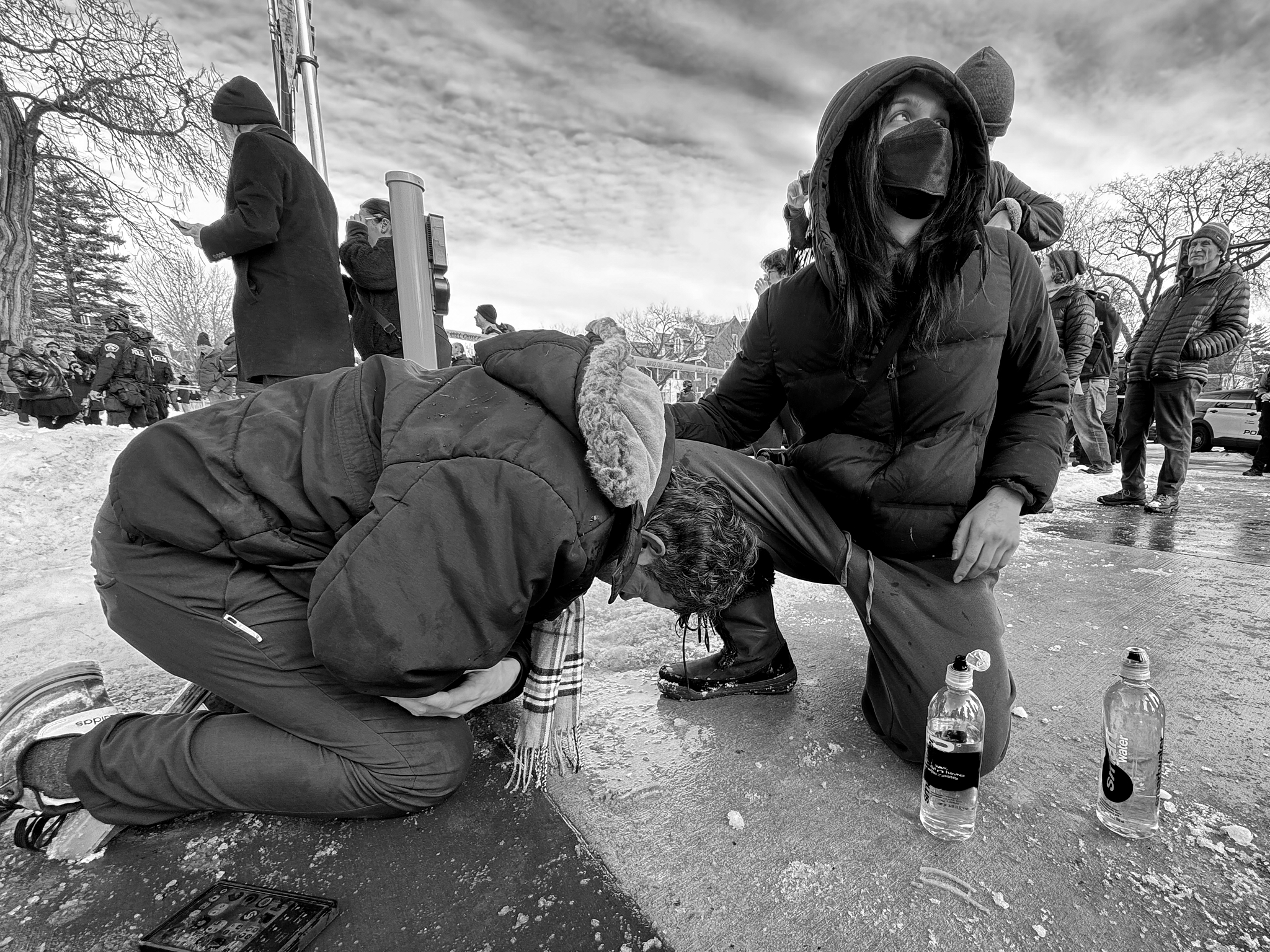 A person collapses to the pavement after being exposed to tear gas during an ICE enforcement action in Minneapolis. Another bystander kneels to help, using water to ease the effects as chemical agents drifted through a residential intersection.