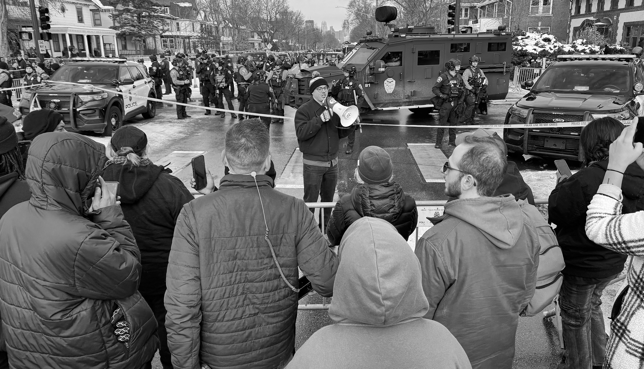 A Minneapolis City Council member, who himself lost an eye after being struck by a police-fired rubber bullet during an earlier protest, addresses demonstrators through a megaphone. He urges them to remain as witnesses as police lines and armored vehicles hold the intersection.