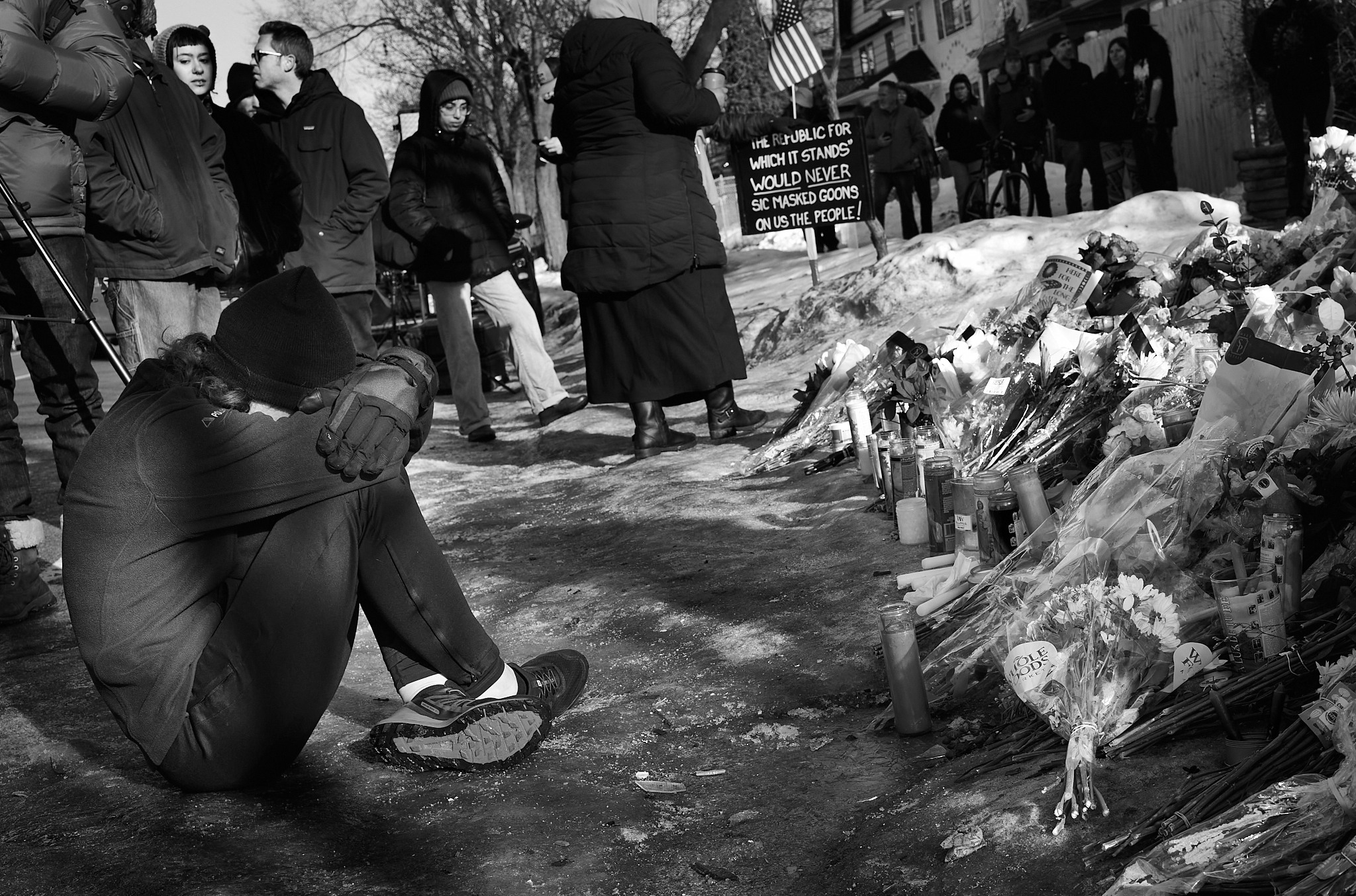 A woman sits beside the memorial, audibly weeping. Around her, the gathering pauses, momentarily still, as her grief fills the space.