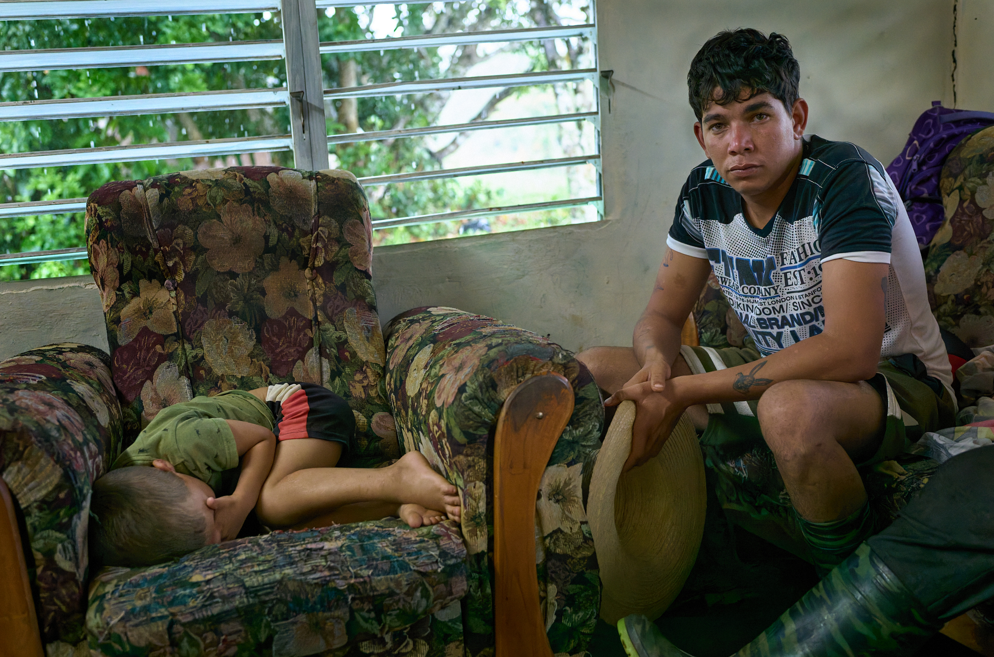 Michael sits beside his younger brother in the living room of their family’s small home. Steel-slatted storm windows let in breeze and light with no screens or air conditioning. Inside, curtain doors and 7-foot walls define rooms beneath an exposed metal roof. A single bare lightbulb illuminates the room.